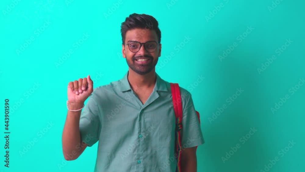 indian student man feeling happy looking at camera wear eye glasses and orange backpack and saying hello with hand in blue studio