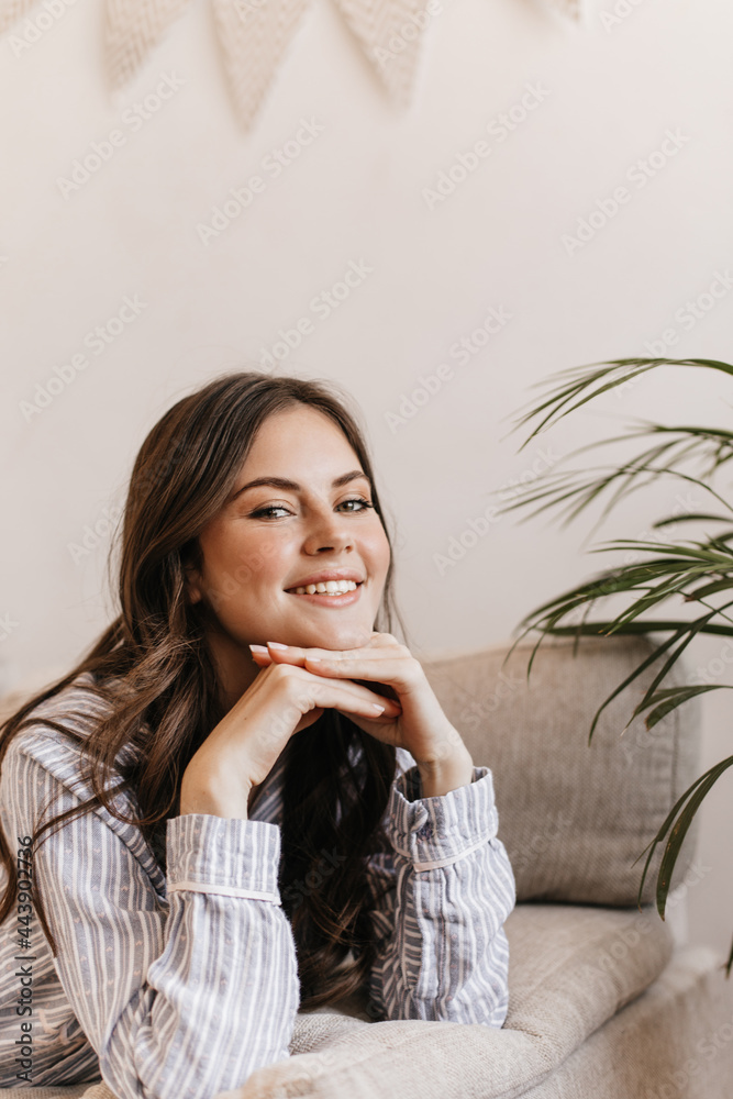 Positive girl in striped outfit leans on sofa in living room. Shot of smiling brunette woman looking into camera and posing at home