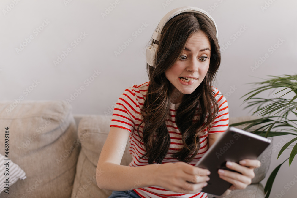 Pretty girls with dark hair are sitting on couch and listening to music in new headphone. Emotional woman in striped t-shirt playing games on her tablet Pretty girls with dark hair are sitting on couch and listening to music in new headphone. Emotional woman in striped t-shirt playing games on her tablet