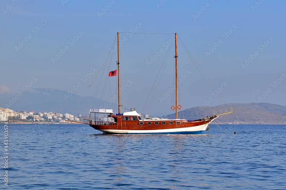 Sailboat with lowered sails under the Albanian flag in bay against background of blue sea and sky