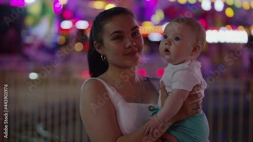 A young mother with a baby in her arms. The kid sees the rides for the first time where there are a lot of flashing lights. Emotions of surprise on the face of an infant