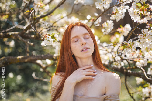 Charming sensual elegant lady with natural red hair touch skin, posing in spring blossom trees. Close up outdoor portrait. Copy, empty space for text