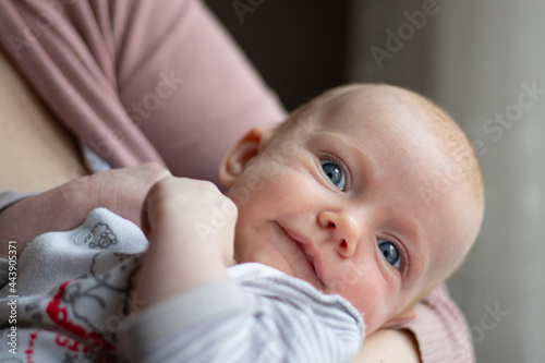 Close-up of a newborn baby with a childish smile, no hair and blue eyes, lying on the mother's arm and looking at the camera. The child is dressed in gray children's clothes. Waist portrait.