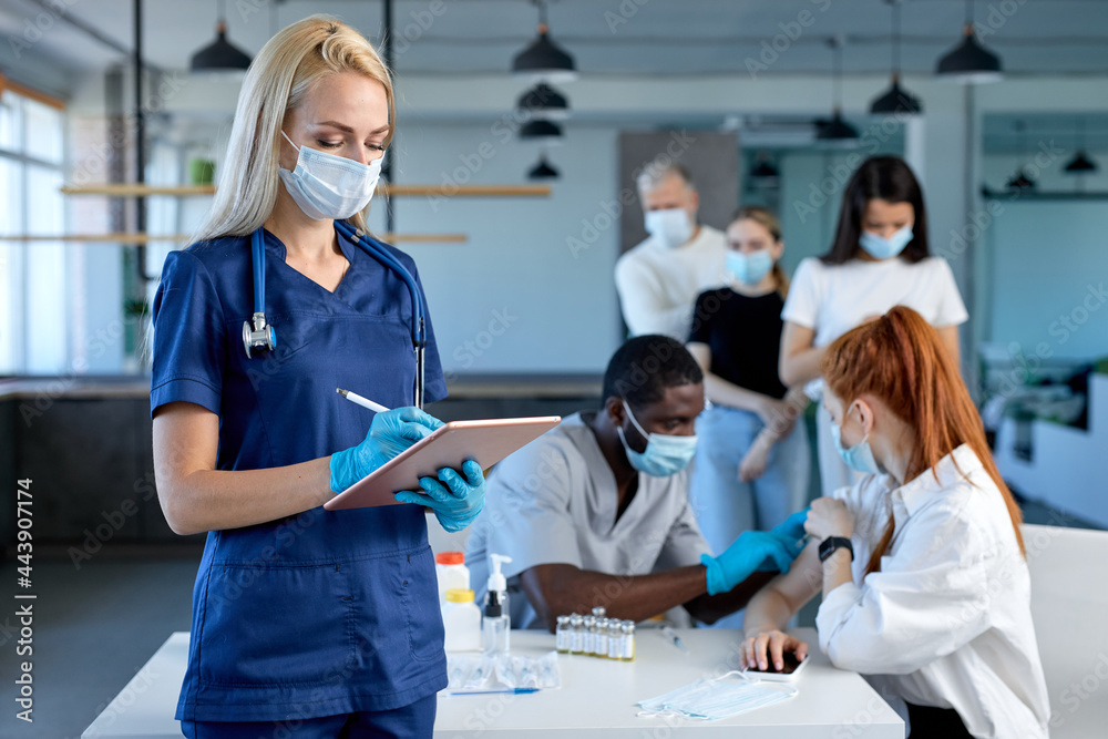 Female doctor at work, beginning of worldwide mass vaccination for coronavirus COVID-19, influenza or flu, world immunization concept. Portrait of blonde nurse in blue uniform holding tablet, writing Female doctor at work, beginning of worldwide mass vaccination for coronavirus COVID-19, influenza or flu, world immunization concept. Portrait of blonde nurse in blue uniform holding tablet, writing