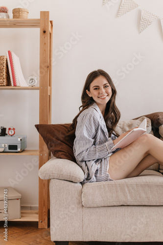 Beautiful girl in striped home clothes with smile looks into camera, holds notebook and rests in living room. Lovely teen in striped shirt posing on beige sofa at home