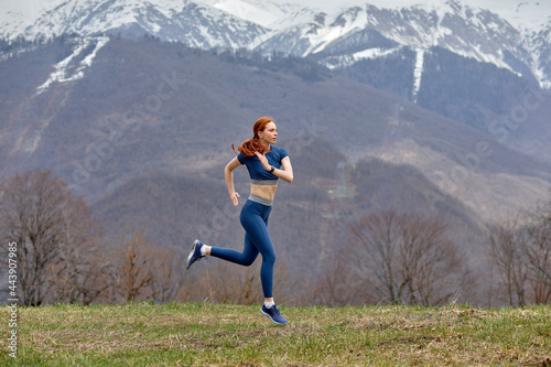 Young woman running outdoors in nature in mountians. Fitness and healthy lifestyle concept. Flexible redhead caucasian lady in blue sportswear lead active lifestyle. Jogging on fresh air
