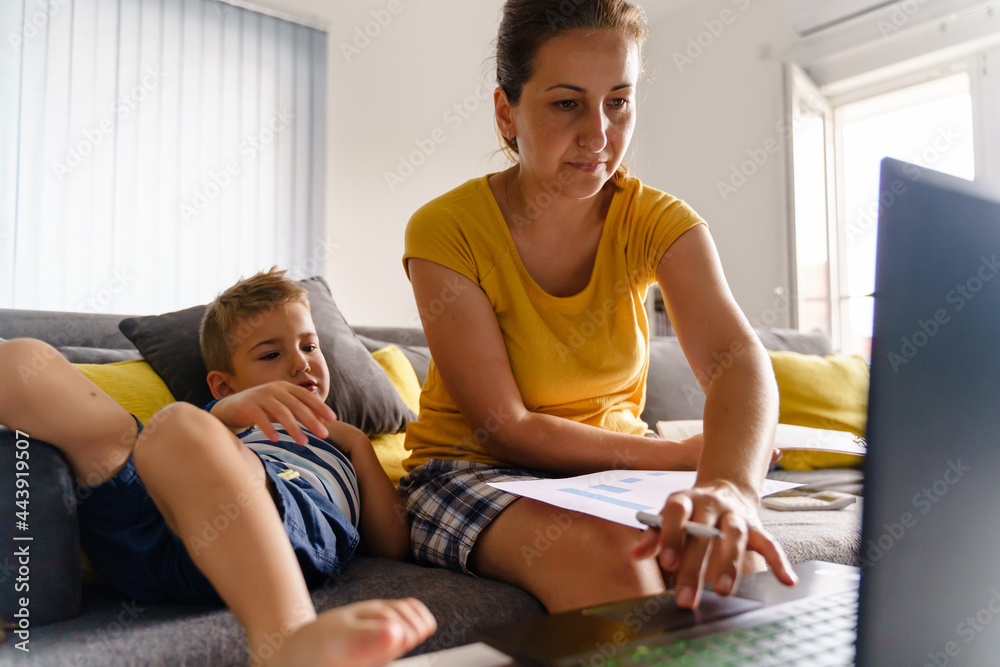 Mature caucasian woman sitting on the sofa at home working on documents while her son child boy is lying on bed beside - work from home entrepreneur female - paperwork preparation business concept Mature caucasian woman sitting on the sofa at home working on documents while her son child boy is lying on bed beside - work from home entrepreneur female - paperwork preparation business concept