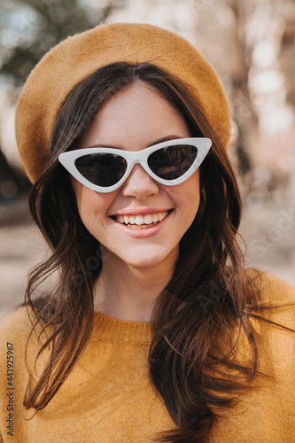 Closeup portrait of smiling girl in yellow beret and sunglasses. Brunette young woman in orange sweater laughing while walking
