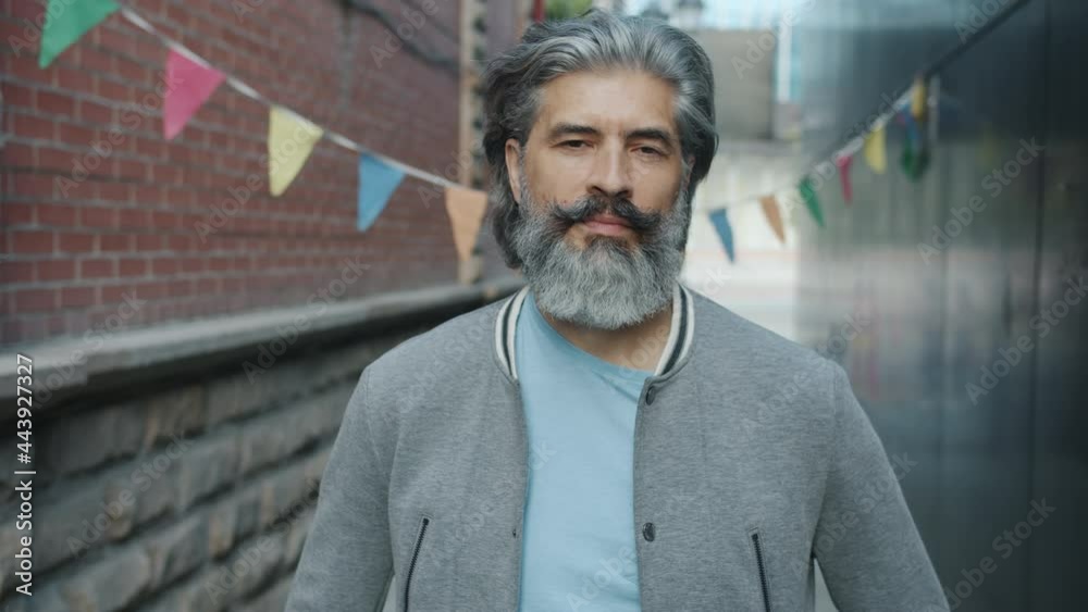 Portrait of pensive guy in casual clothing standing outdoors against urban background and looking at camera with calm and serious face. People and emotions concept.