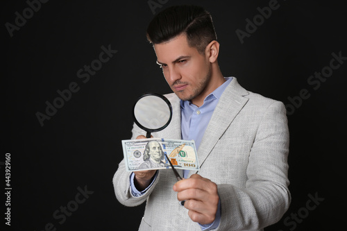 Expert authenticating 100 dollar banknote with magnifying glass against black background. Fake money concept photo