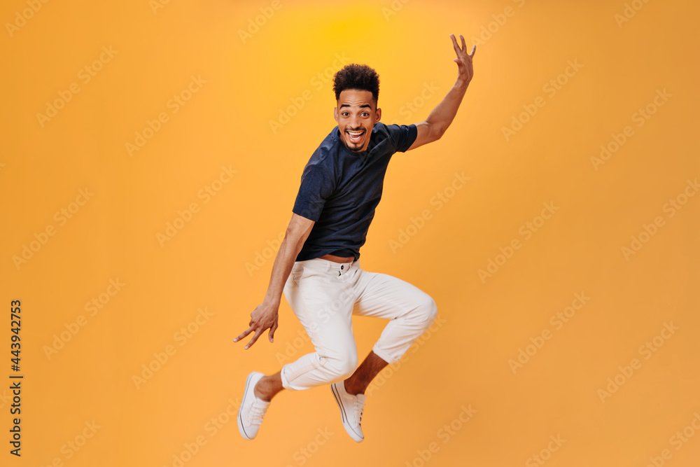 Cheerful guy in black T-shirt jumping high on orange background. Snapshot of man in white pants dancing and having fun on isolated