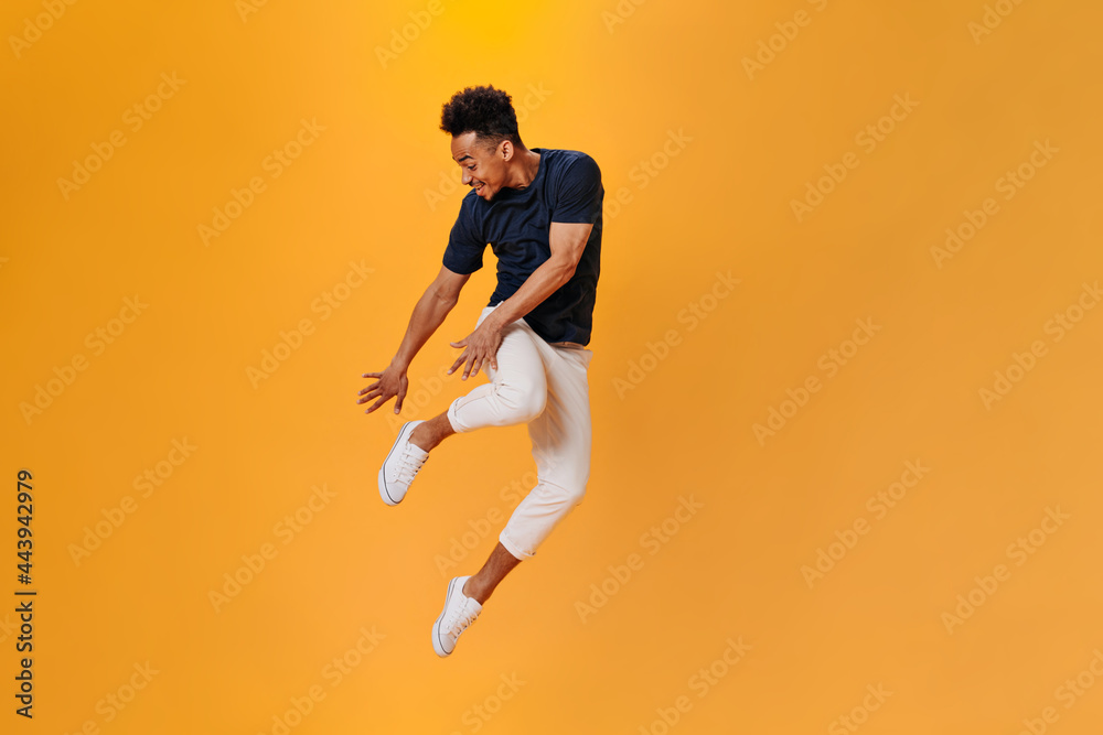 Young man in white pants and black T-shirt jumping high on orange background. Brunette guy having fun on isolated backdrop Young man in white pants and black T-shirt jumping high on orange background. Brunette guy having fun on isolated backdrop