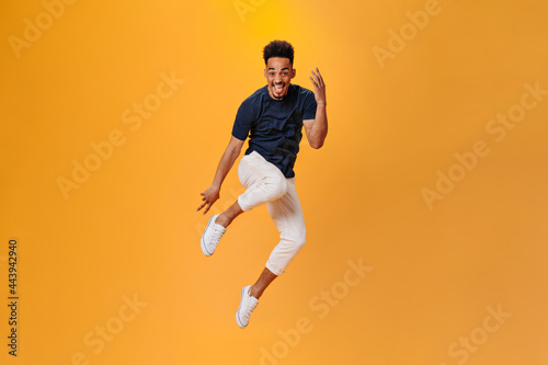 Optimistic dark skinned guy in stylish outfit jumping on orange background. Brunette man in black t-shirt and white pants posing on isolated