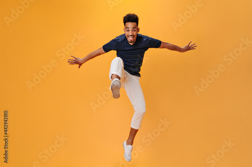 Handsome man in black t-shirt smiles and jumps on orange background. Cool guy in white pants moving on isolated backdrop
