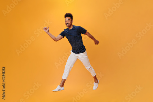 Joyful young man walks on orange background. Cool dark-haired guy in white pants and tee jumping on isolated backdrop