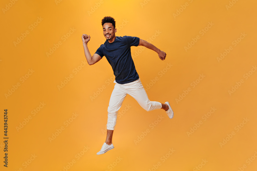 Perky man in light pants jumping and having fun on orange background. Snapshot of brunette guy in white trousers posing on isolated