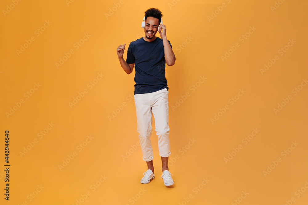 Fototapeta premium Stylish man in white pants listens to music in massive headphones. Portrait of guy in dark-blue t-shirt dancing on orange background