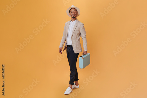 Handsome man in hat and suit posing with suitcase on orange background. Young guy in beige jacket and black pants holding blue bag