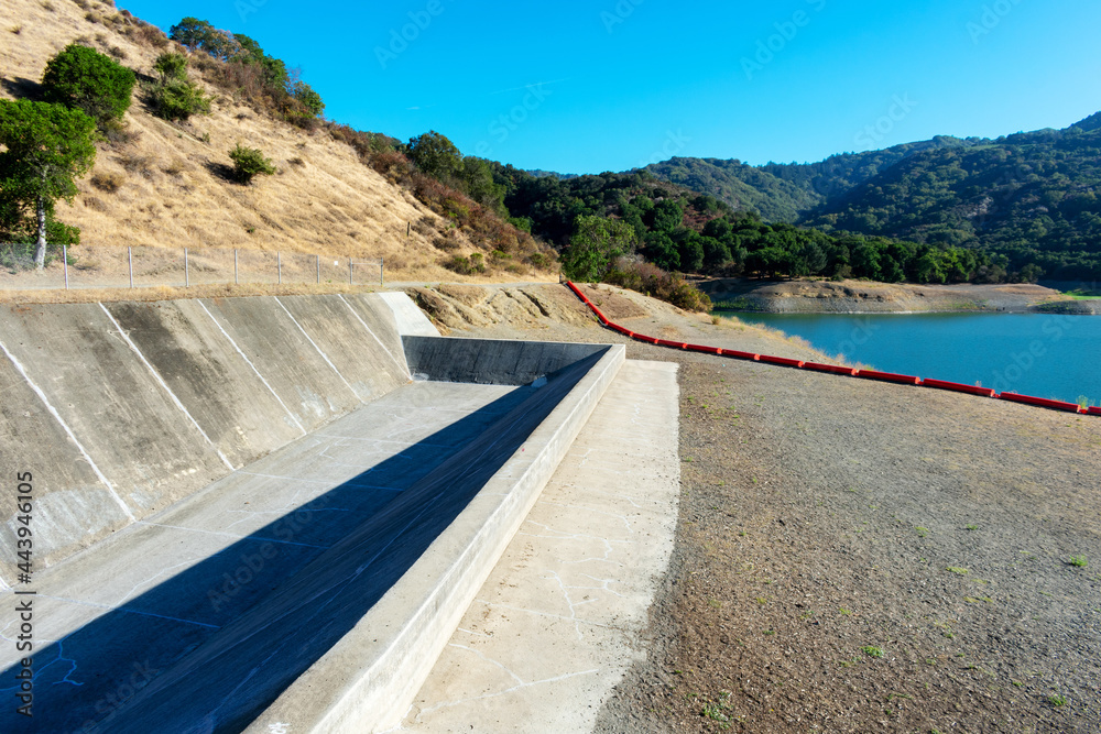 Concrete dam spillway, orange debris boom on dry ground. Extremely low water level in drying during summer Stevens Creek reservoir in San Francisco Bay Area, California