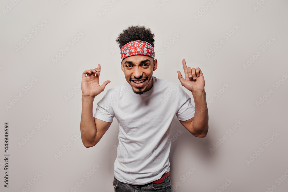 Fototapeta premium Cool guy in bandana looks into camera on white background. Portrait of man in white t-shirt and black jeans smiling and dancing on isolated
