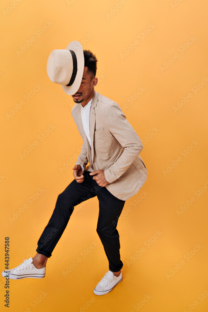 Guy in beige jacket and black pants posing on orange background. Portrait of man in suit and hat dancing on isolated backdrop Guy in beige jacket and black pants posing on orange background. Portrait of man in suit and hat dancing on isolated backdrop