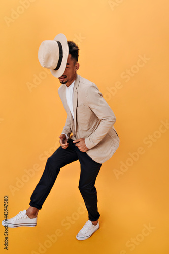 Guy in beige jacket and black pants posing on orange background. Portrait of man in suit and hat dancing on isolated backdrop