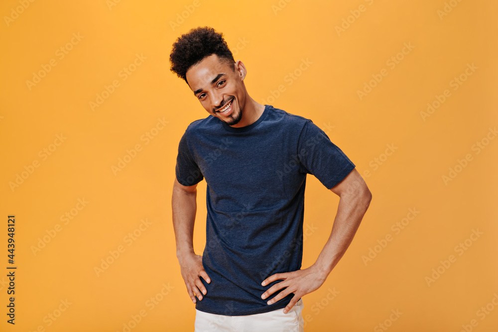 Stylish cute guy smiling and looking at camera on orange background. Attractive brunette man in blue t-shirt posing on isolated backdrop Stylish cute guy smiling and looking at camera on orange background. Attractive brunette man in blue t-shirt posing on isolated backdrop