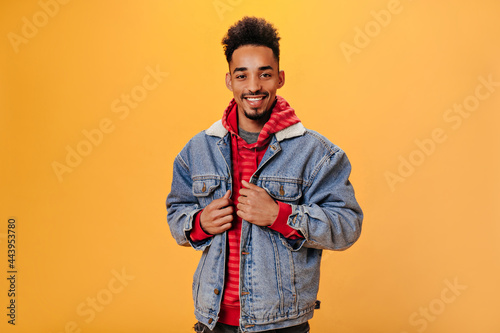 Dark-skinned guy in stylish outfit with smile posing on orange background. Portrait of brunette man in denim jacket looking into camera on isolated