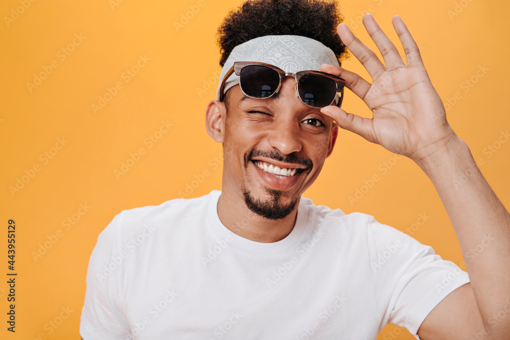 Man in stylish outfit removes glasses and winks. Brunette dark eyed guy in white t-shirt widely smiling on orange isolated backgound Man in stylish outfit removes glasses and winks. Brunette dark eyed guy in white t-shirt widely smiling on orange isolated backgound