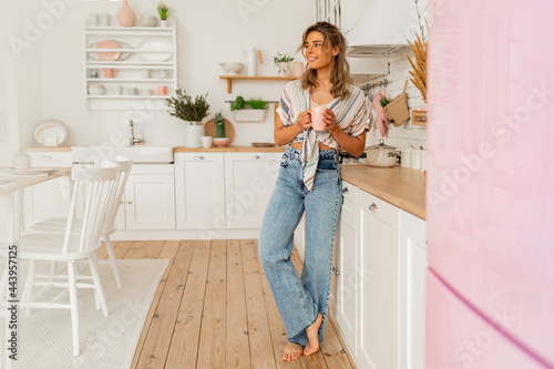 Pretty woman enjoying cozy morning on her stylish kitchen while drinking hot tea. Full length.