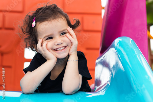 a cute little girl laying on the blue slide in a playground with hands under her chin.
