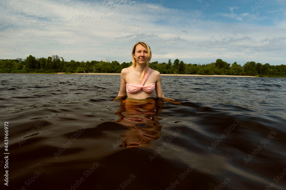 Fototapeta premium Portrait of wet blonde woman in swimsuit in dark water on nature. Swimming on the river