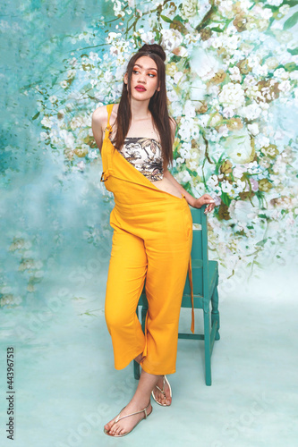 Beautiful female mixed race model with pale skin, in a floral studio setting, wearing a yellow jumpsuit and using a chair to pose.  © Anina Lonte