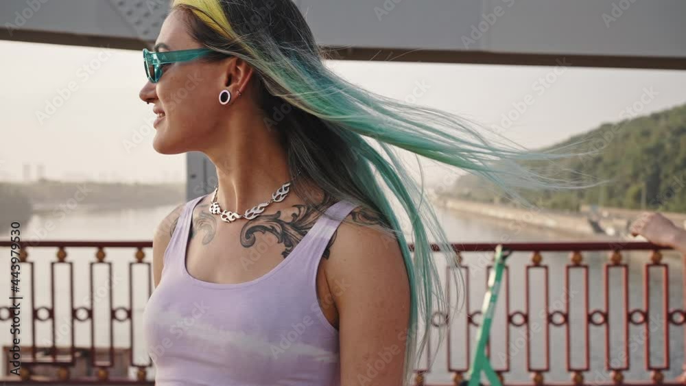 Active girl with bright hair and a tattoo is riding a skateboard across the bridge in the company of her friends, skateboarders. Youth culture, urban landscape