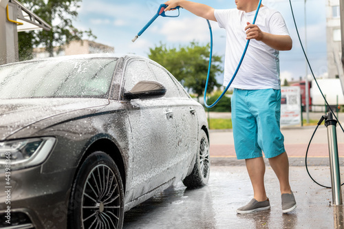 Close-up detail car wash with high pressure water equipment pump at self-service outdoor on bright shiny summer day. Vehicle covered with foam shampoo chemical detergents during carwash self service photo