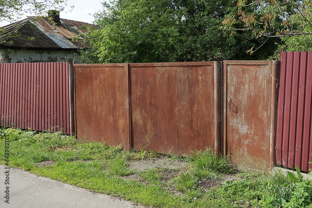 one closed rusty iron gate with a closed door and part of a red metal fence on a rural street in green grass one closed rusty iron gate with a closed door and part of a red metal fence on a rural street in green grass