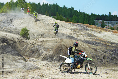 Four motorcyclists climb up a clay slope. The fifth biker stayed downstairs and watched the departing motorcyclists. All bikers are dressed in bright clothes and helmets.