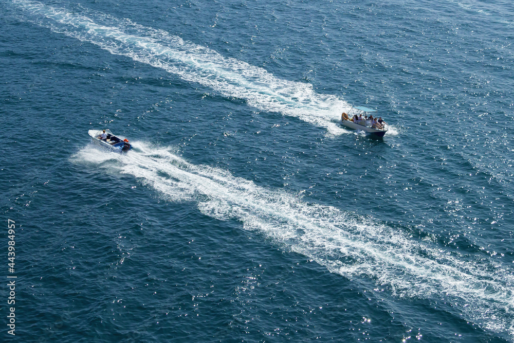Obraz premium Two white and blue boats sail in different directions on the blue sea. White, foamy footprints follow the boats. The direction of movement of vessels is opposite. Boats is carrying a group of turists.