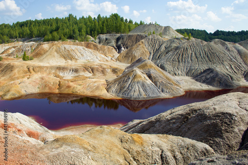 A dark red, acidic river against the backdrop of clay hills, a blue sky with clouds and a green forest. Hills, sky and forest are reflected in the surface of the water.