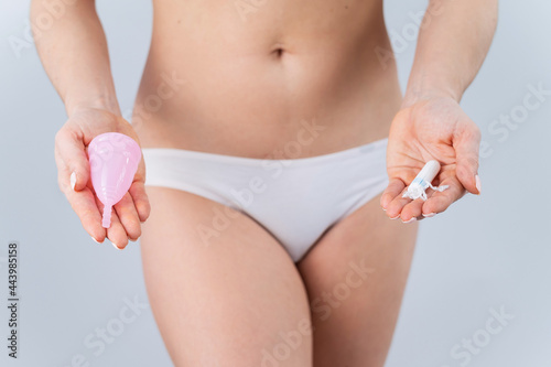 A faceless woman in cotton panties holds a pink menstrual cup and tampon on a white background. Various hygiene products during menstruation
