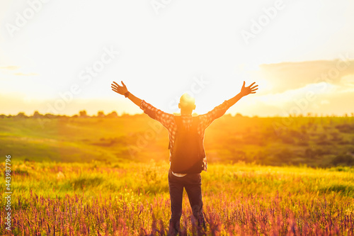 Fototapeta Naklejka Na Ścianę i Meble -  Traveler with raised hands standing on a hill in green grass and enjoying a majestic sunset, Successful man enjoying triumph
