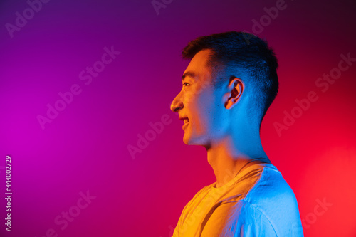 Side view. Asian young man isolated on studio background in gradient pink purple neon light, colour filter. Concept of human emotions, facial expression.