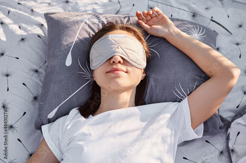 Outdoor shot of young brunette female wearing white casual style t shirt and sleeping mask lying on bed and sleeping, enjoying fresh air and sunshine.