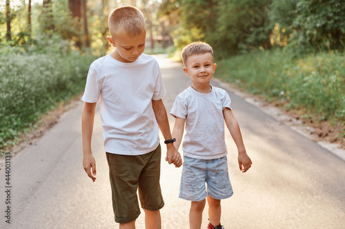 Two little boys wearing white t shirts and shorts going together and holding hands in summer park, brothers walking outdoor, expressing positive emotions.