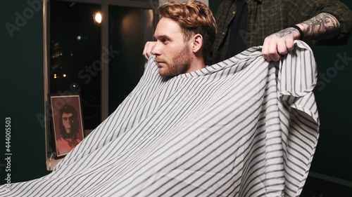 A cropped view of a skillful barber is putting on an apron on his client before cutting hair in a barber shop