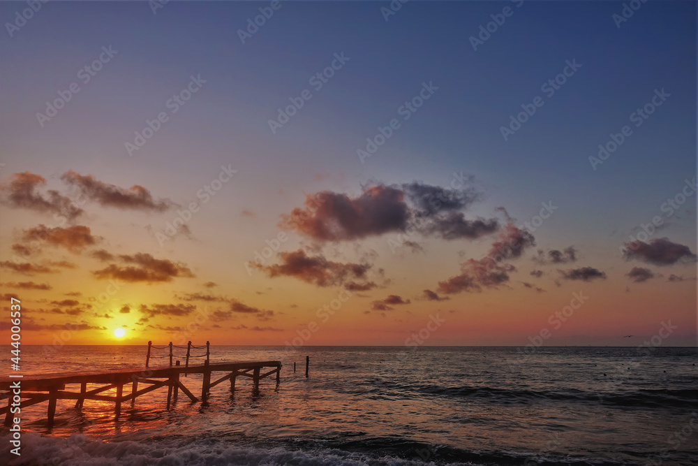 Bright sunrise over the Caribbean Sea. The sun is low on the horizon. The sky is tinted with golden and scarlet hues. Lilac clouds. There is a wooden footpath over the water. Foam waves on the beach.