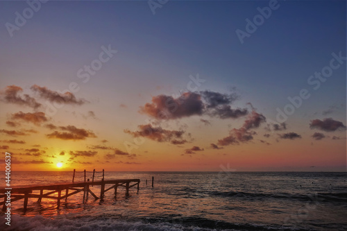 Bright sunrise over the Caribbean Sea. The sun is low on the horizon. The sky is tinted with golden and scarlet hues. Lilac clouds. There is a wooden footpath over the water. Foam waves on the beach.