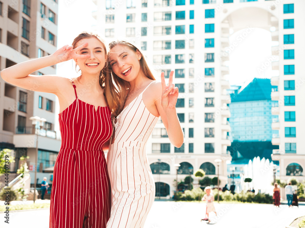 Two young beautiful smiling hipster female in trendy overalls.Sexy carefree women posing on the street background. Positive models having fun, hugging and going crazy.Show peace sign