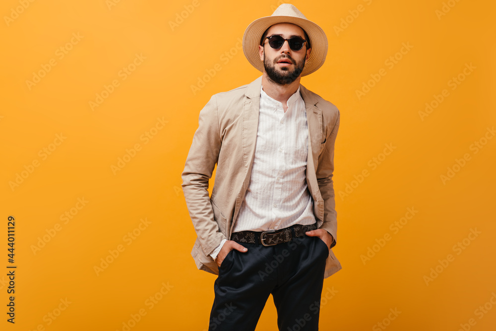 Stylish guy in beige jacket and wide brimmed hat posing on orange background