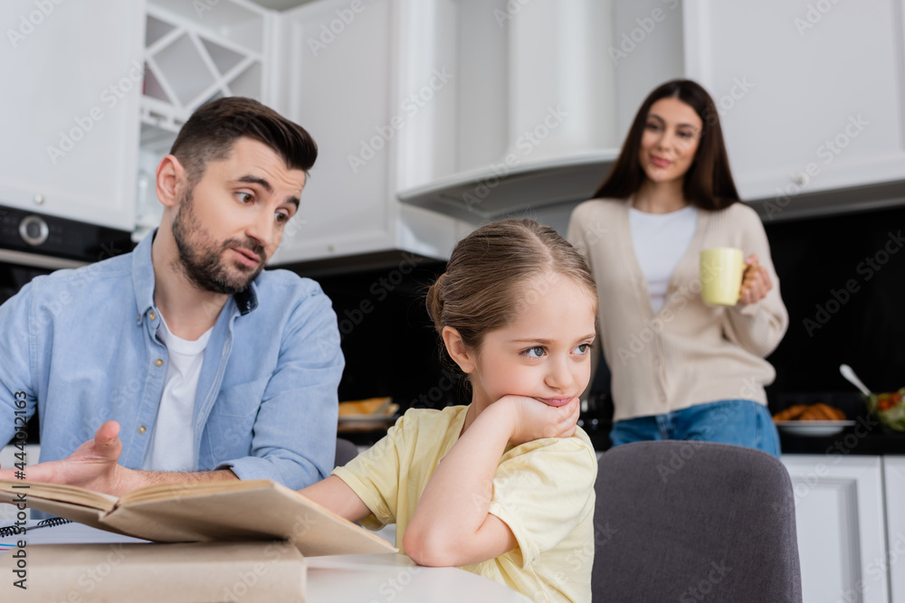man talking to offended daughter while doing homework near wife on blurred background
